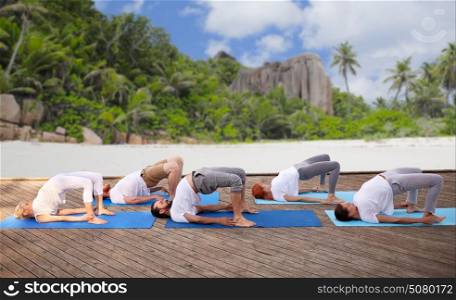 fitness, sport, yoga and healthy lifestyle concept - group of people making bridge pose over tropical beach background. group of people making yoga exercises over beach