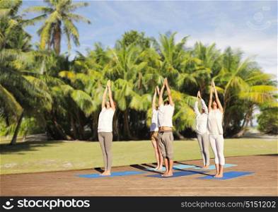 fitness, sport, yoga and healthy lifestyle concept - group of people making upward salute pose over natural exotic background with palm trees. group of people making yoga exercises outdoors