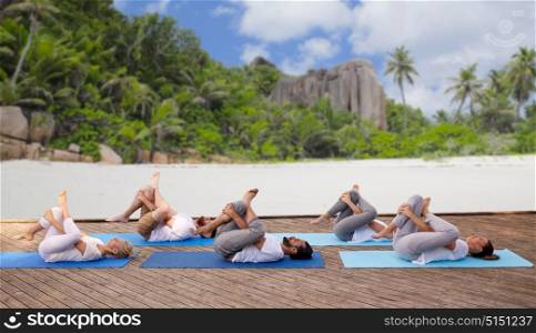 fitness, sport, yoga and healthy lifestyle concept - group of people making half ankle to knee supine pose over tropical beach background. group of people making yoga exercises over beach