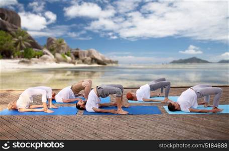 fitness, sport, yoga and healthy lifestyle concept - group of people making bridge pose over tropical beach background. group of people making yoga exercises over beach
