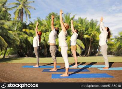 fitness, sport, yoga and healthy lifestyle concept - group of people making upward salute pose over natural exotic background with palm trees. group of people making yoga exercises outdoors