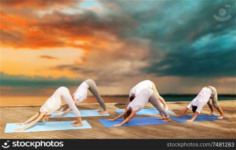 fitness, sport, yoga and healthy lifestyle concept - group of people making downward facing dog pose on sea pier over sunset background. group of people making yoga dog pose outdoors