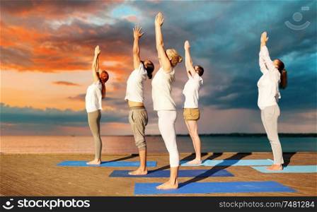 fitness, sport, yoga and healthy lifestyle concept - group of people making upward salute pose on sea pier over sunset background. group of people making yoga exercises outdoors