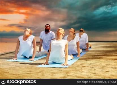 fitness, sport, yoga and healthy lifestyle concept - group of people making cobra pose on sea pier over sunset background. group of people making yoga exercises outdoors