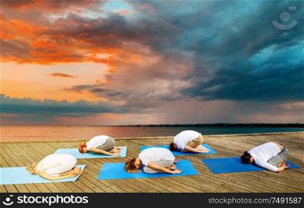 fitness, sport, yoga and healthy lifestyle concept - group of people making child pose on sea pier over sunset background. group of people making yoga exercises outdoors
