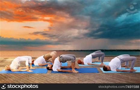 fitness, sport, yoga and healthy lifestyle concept - group of people making bridge pose on sea pier over sunset background. group of people making yoga exercises outdoors