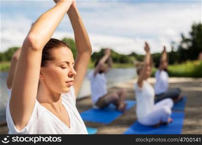 fitness, sport, yoga and healthy lifestyle concept - group of people meditating in lotus pose on river or lake berth
