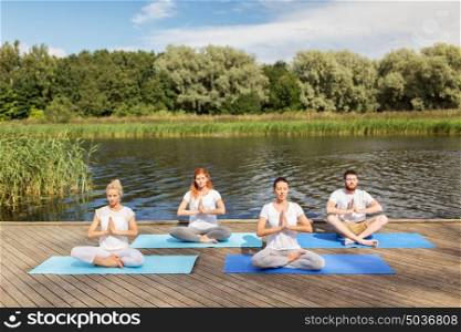 fitness, sport, yoga and healthy lifestyle concept - group of people meditating in lotus pose on river or lake berth. people meditating in yoga lotus pose outdoors