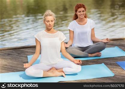 fitness, sport, yoga and healthy lifestyle concept - women meditating in lotus pose on river or lake berth. women meditating in yoga lotus pose outdoors