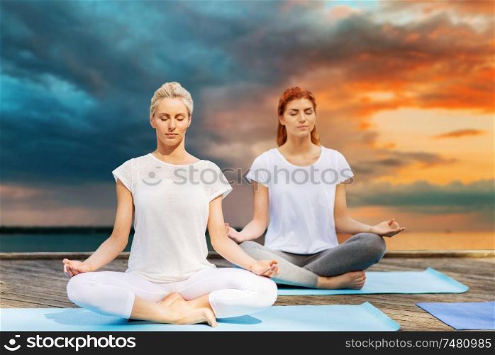 fitness, sport, yoga and healthy lifestyle concept - women meditating in lotus pose on sea pier over sunset background. women meditating in yoga lotus pose outdoors