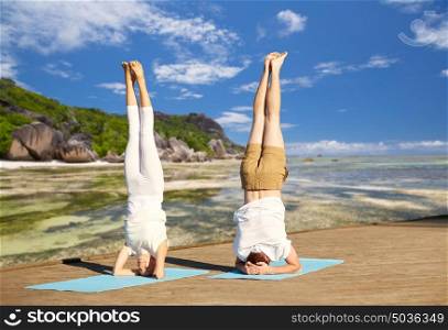 fitness, sport, yoga and people concept - couple making headstand pose on mat over exotic tropical beach background. couple making yoga headstand on mat outdoors