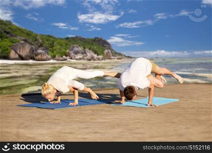fitness, sport, yoga and people concept - couple making side crane pose on mat over exotic tropical beach background. couple making yoga side crane pose outdoors