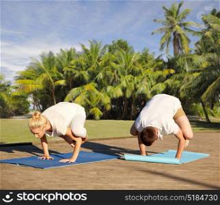 fitness, sport, yoga and people concept - couple making side crow pose on mat over natural background with palm trees . couple making yoga crow pose outdoors