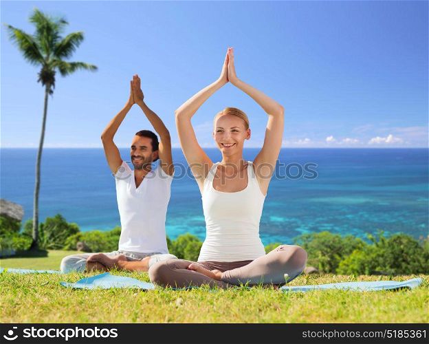 fitness, sport, yoga and people concept - happy couple meditating in lotus pose outdoors over exotic natural background with palm tree and ocean. couple doing yoga in lotus pose outdoors