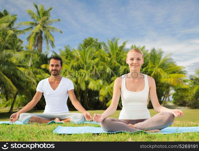 fitness, sport, yoga and people concept - happy couple meditating in lotus pose outdoors over exotic natural background with palm trees. couple doing yoga in lotus pose outdoors