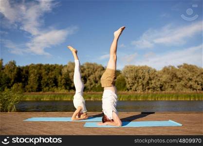 fitness, sport, yoga, people and healthy lifestyle concept - couple making headstand pose on mat on river or lake berth