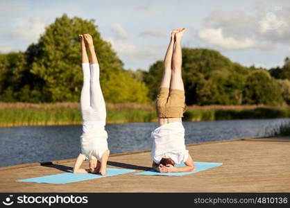 fitness, sport, yoga, people and healthy lifestyle concept - couple making headstand pose on mat on river or lake berth