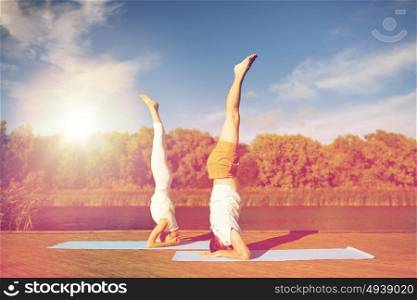 fitness, sport, yoga, people and healthy lifestyle concept - couple making headstand pose on mat on river or lake berth. couple making yoga headstand on mat outdoors