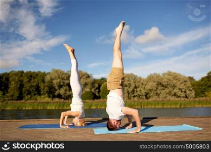 fitness, sport, yoga, people and healthy lifestyle concept - couple making headstand pose on mat on river or lake berth. couple making yoga headstand on mat outdoors