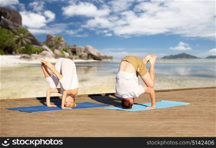fitness, sport, yoga, people and healthy lifestyle concept - couple making headstand outdoors over tropical beach background. couple making yoga over tropical beach background