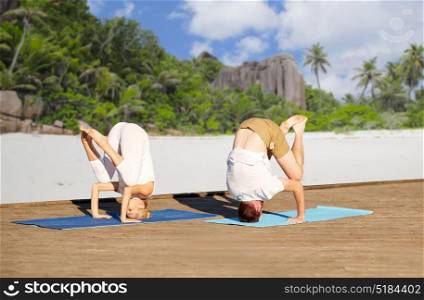 fitness, sport, yoga, people and healthy lifestyle concept - couple making headstand outdoors over tropical beach background. couple making yoga over tropical beach