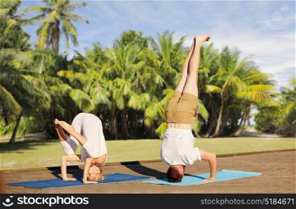 fitness, sport, yoga, people and healthy lifestyle concept - couple making headstand outdoors over natural exotic background with palm trees. couple making yoga outdoors