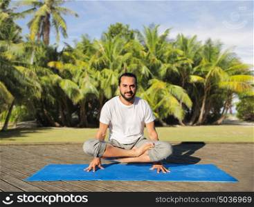 fitness, sport, yoga, people and healthy lifestyle concept - man making scale pose lotus variation on mat outdoors over exotic natural background with palm trees. man making yoga in scale pose outdoors