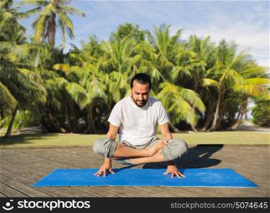 fitness, sport, yoga, people and healthy lifestyle concept - man making scale pose lotus variation on mat outdoors over exotic natural background with palm trees. man making yoga in scale pose outdoors