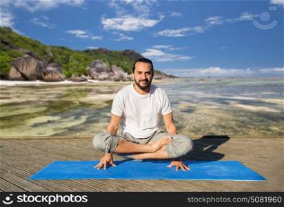 fitness, sport, yoga, people and healthy lifestyle concept - man making scale pose lotus variation on mat over exotic tropical beach background. man making yoga in scale pose outdoors
