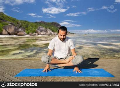 fitness, sport, yoga, people and healthy lifestyle concept - man making scale pose lotus variation on mat over exotic tropical beach background. man making yoga in scale pose outdoors