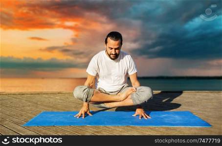 fitness, sport, yoga, people and healthy lifestyle concept - man making scale pose lotus variation on mat outdoors on sea pier over sunset background. man making yoga in scale pose outdoors