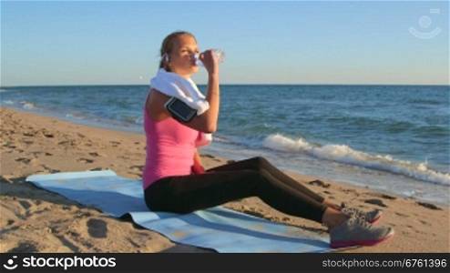 Fitness sporty woman in wireless headset relaxing after workout listening music and drinking water on the beach