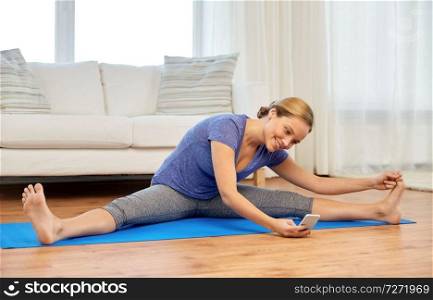 fitness, technology and gymnastics concept - woman with smartphone doing yoga or split exercise at home. woman with smartphone doing yoga at home