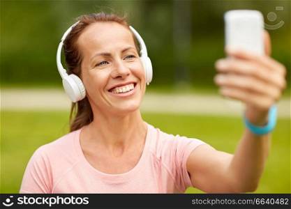 fitness, technology and healthy lifestyle concept - smiling woman in headphones listening to music and taking selfie by smartphone at summer park. woman in phones takes selfie by smartphone at park