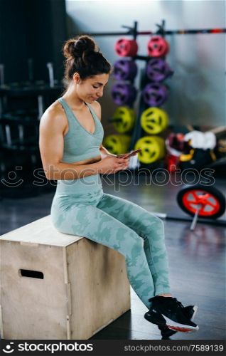 Fitness woman consulting her training on her smartphone sitting in a jump box in the gym. Woman consulting her training on her smartphone sitting in a jump box