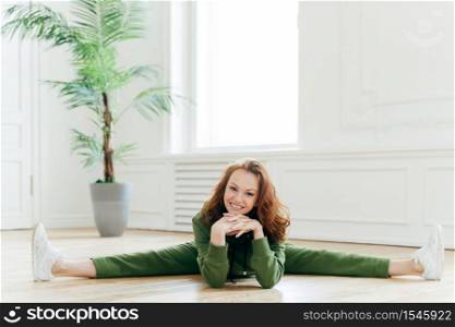 Fitness woman demonstrates nice flexibility, does gymnastics exercises, shows leg split, keeps hands under chin, smiles broadly, poses on floor in spacious room. People, workout, aerobics concept