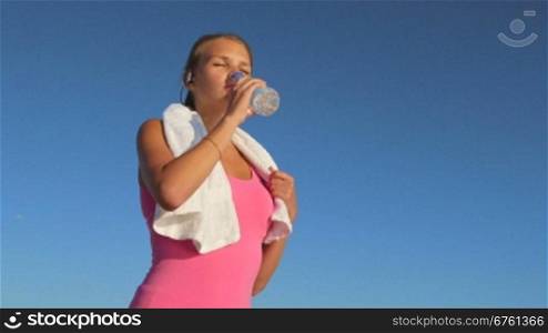 Fitness woman drinking water after exercising on summer day against blue sky