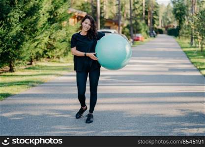 Fitness woman exercises with fitness ball outdoor walks on road near forest has healthy lifestyle dressed in black active wear, has happy expression, stays in good physical shape. Fitness concept