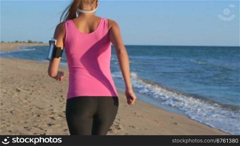 Fitness woman jogging on beach listening music in earphones from smart phone mp player rear view