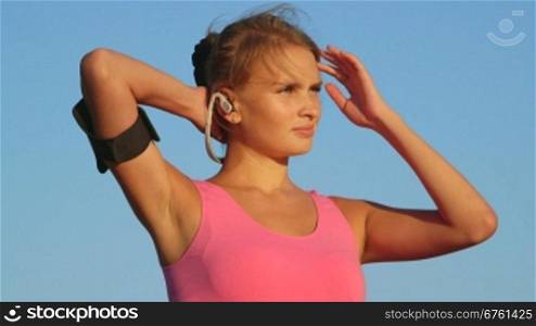 Fitness woman on workout with earphones and sport smartphone armband looking away against blue sky