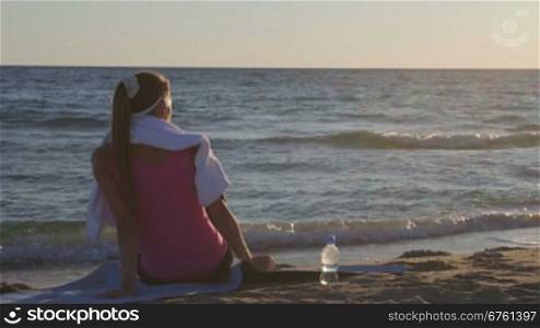 Fitness woman relaxing after workout routine listening music on beach at sunset rear view