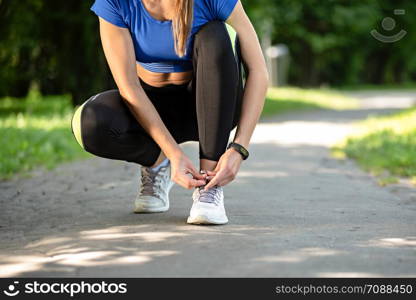 Fitness woman tying laces in running shoes during outdoors workout in the park on a sunny morning