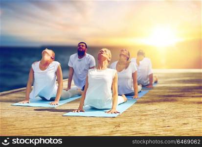 fitness, yoga and healthy lifestyle concept - group of people doing cobra pose outdoors on wooden pier over sea background. group of people doing cobra pose outdoors