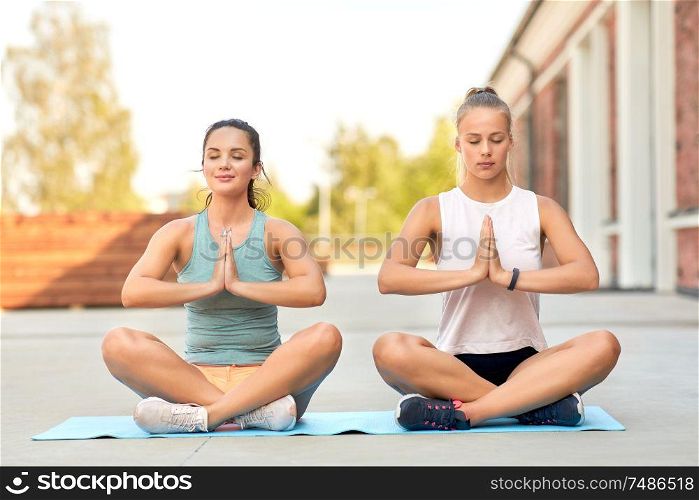 fitness, yoga and healthy lifestyle concept - young women or female friends meditating in lotus pose on mat outdoors. women doing yoga and meditating in lotus pose