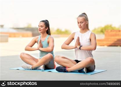 fitness, yoga and healthy lifestyle concept - young women or female friends meditating in lotus pose on mat outdoors. women doing yoga and meditating in lotus pose