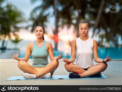 fitness, yoga and healthy lifestyle concept - young women or female friends meditating in lotus pose on mat over tropical beach on background. women doing yoga and meditating in lotus pose