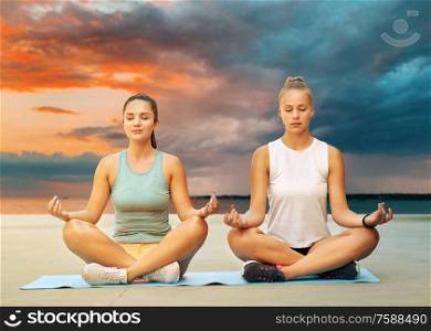 fitness, yoga and healthy lifestyle concept - young women or female friends meditating in lotus pose on mat over sea and sunset sky on background. women doing yoga and meditating in lotus pose