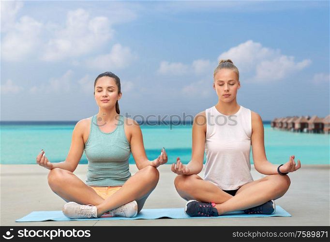 fitness, yoga and healthy lifestyle concept - young women or female friends meditating in lotus pose on mat over sea and bungalow on background. women doing yoga and meditating in lotus pose