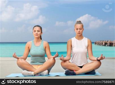 fitness, yoga and healthy lifestyle concept - young women or female friends meditating in lotus pose on mat over sea and bungalow on background. women doing yoga and meditating in lotus pose