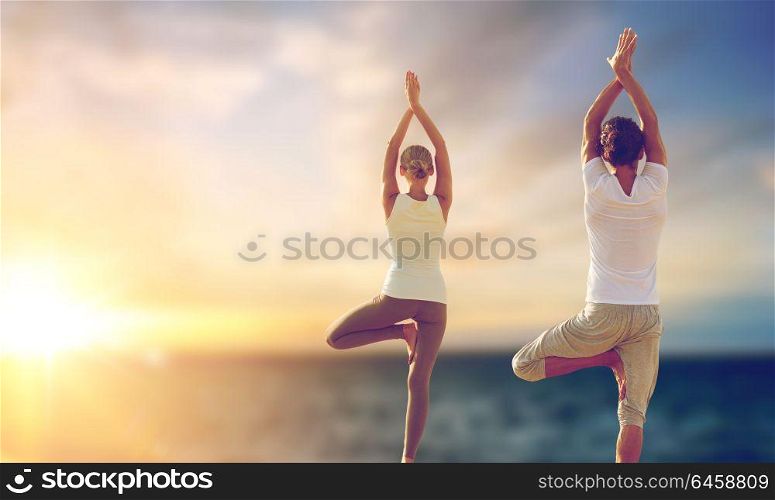 fitness, yoga and people concept - happy couple making tree pose and meditating outdoors over sea background. happy couple making yoga tree pose over sea